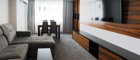 Una habitación de hotel moderna con un sofá modular, un televisor de pantalla plana montado en un panel de madera, un área de comedor con mesa y sillas, y un ventanal con cortinas.