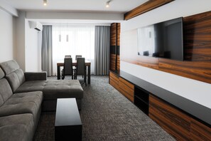 A modern hotel room with a sectional sofa, a flat-screen TV mounted on a wooden panel, a dining area with a table and chairs, and a large window with curtains. - Hotel Sport (Belchatow)