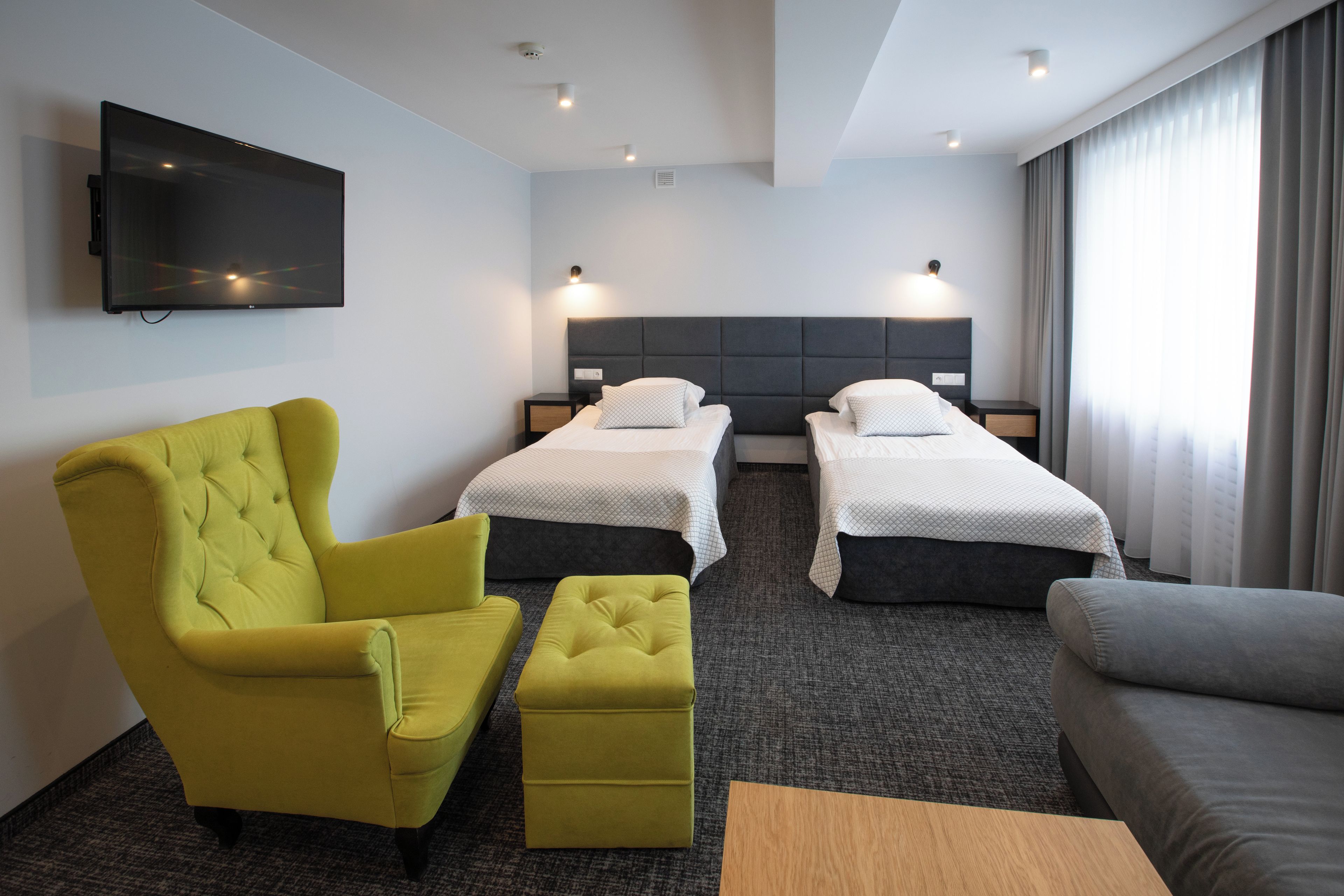 Una habitación de hotel con dos camas individuales, un sillón amarillo, un sofá gris y un televisor de pantalla plana montado en la pared.