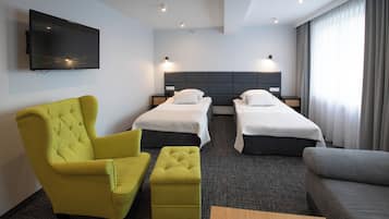 Una habitación de hotel con dos camas individuales, un sillón amarillo, un sofá gris y un televisor de pantalla plana montado en la pared.