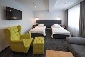 Una habitación de hotel con dos camas individuales, un sillón amarillo, un sofá gris y un televisor de pantalla plana montado en la pared.