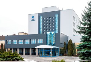 A modern hotel building with a prominent logo, a covered entrance, and a parking area. - Hotel Sport (Belchatow)
