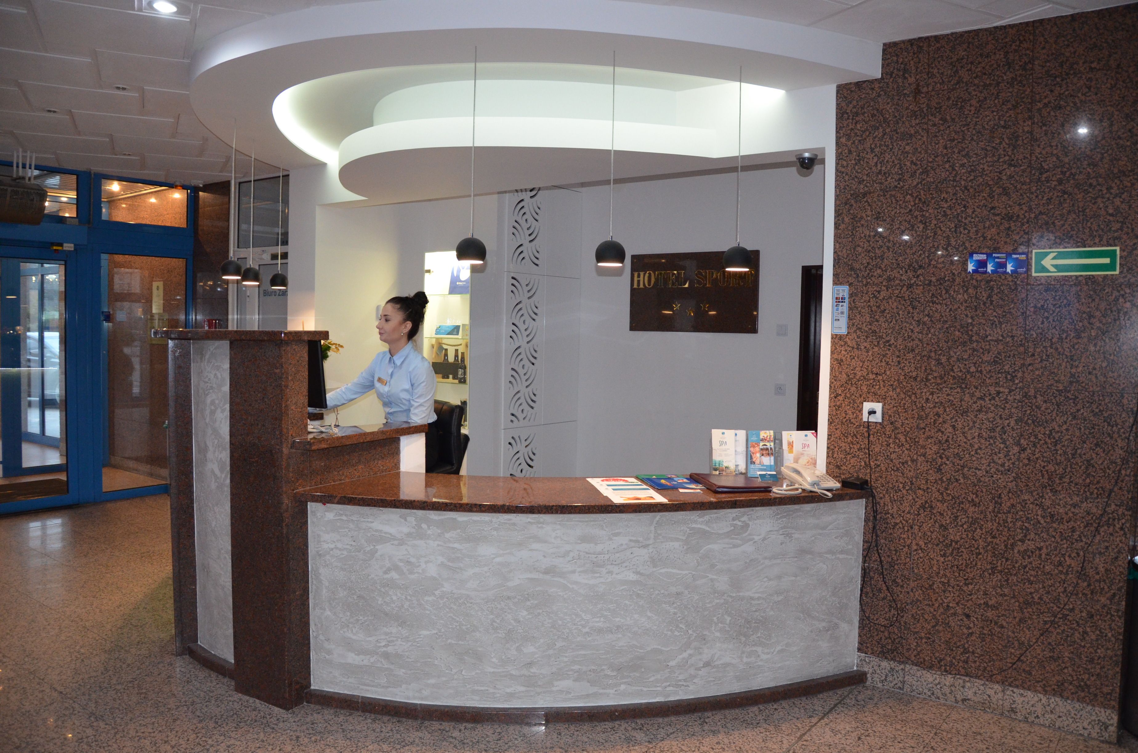 Een receptiebalie van een hotel met een receptionist, een bord met de tekst 'HOTEL SPREE' en een richtingwijzer die naar links wijst.