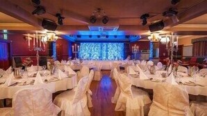 A banquet hall with rows of tables set for a formal event, equipped with chandeliers and a stage at the front. - Lake Of Shadows (Buncrana)