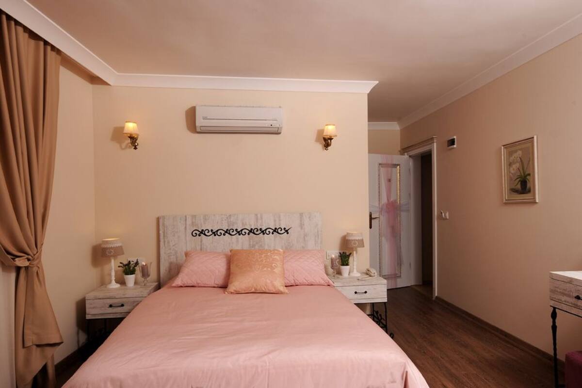 A bedroom with a bed, two bedside tables, an air conditioning unit, and a door leading to another room.