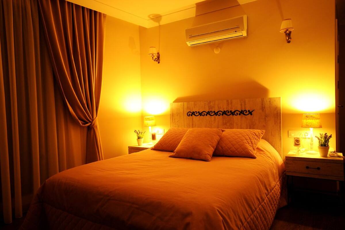 A hotel room with a large bed, two bedside tables with lamps, a wall-mounted air conditioner, and a curtain on the left.