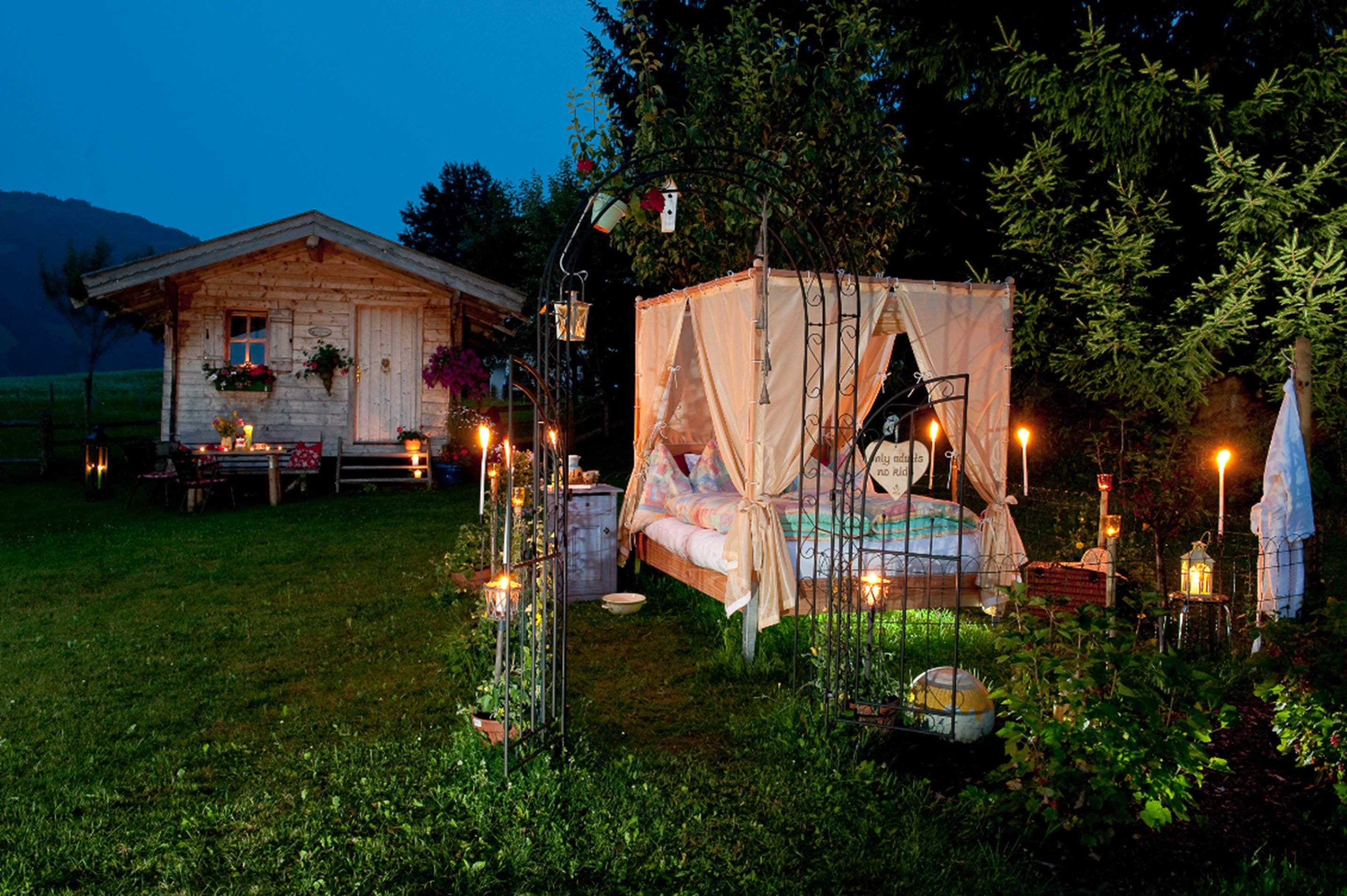 A rustic wooden cabin with a bed and a small table, surrounded by greenery and decorative lights.