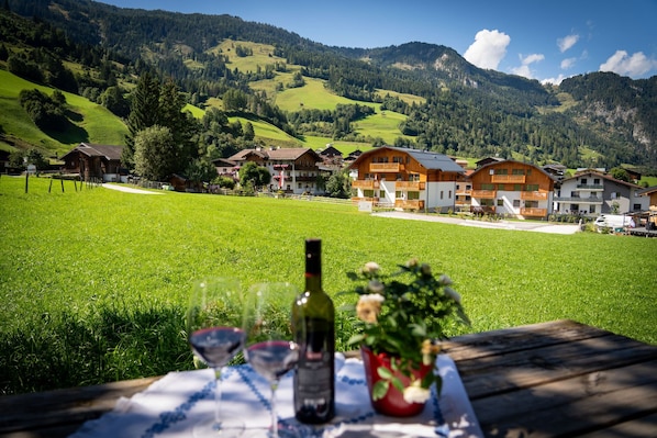 A table with wine glasses and bottles, set against a backdrop of a green field and a row of wooden houses with balconies, under a clear sky. - die Unterbergerin (Dorfgastein)