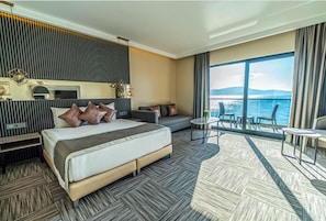 A modern hotel room with a large bed, a sofa, a small table, and a balcony with a view of the sea. - Assos Dove Resort & Spa Hotel (Ayvacik)