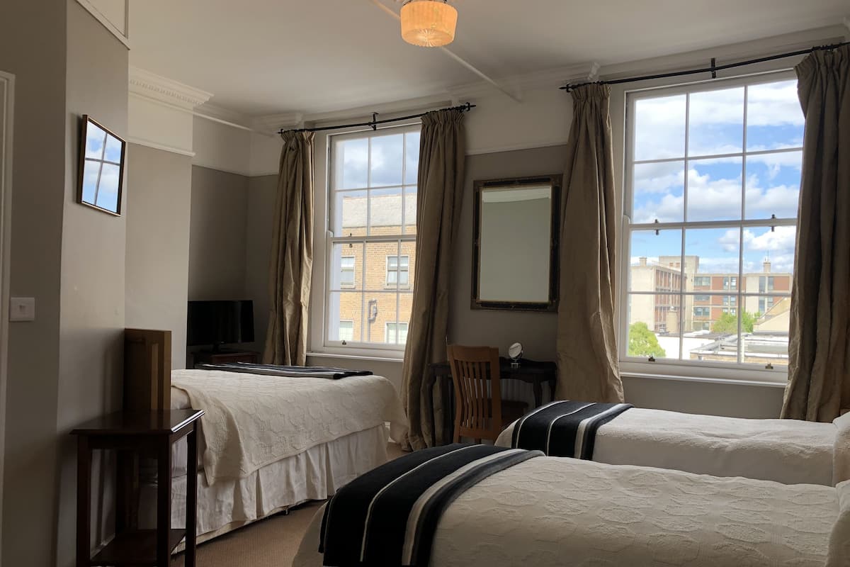 A bedroom with two beds, a desk, a chair, a TV, and large windows with curtains.