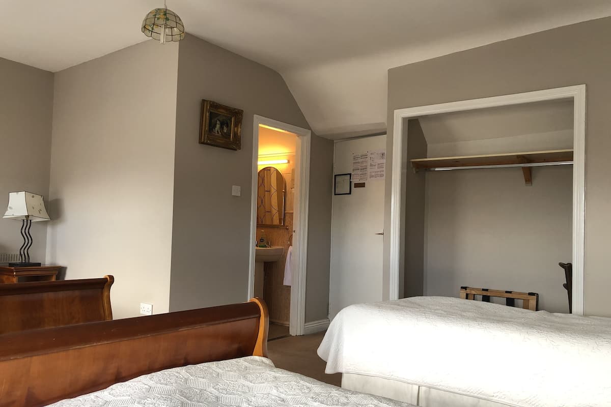 A bedroom with two beds, a wooden headboard, a bedside table with a lamp, a wardrobe, and a bathroom visible through an open door.