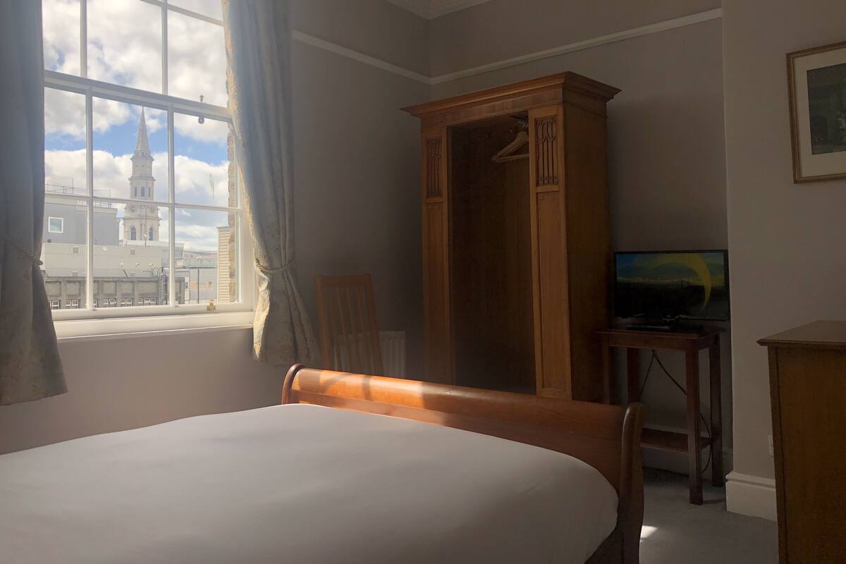 A hotel room with a large bed, a wooden wardrobe, a television on a small table, and a window with a view of a building.