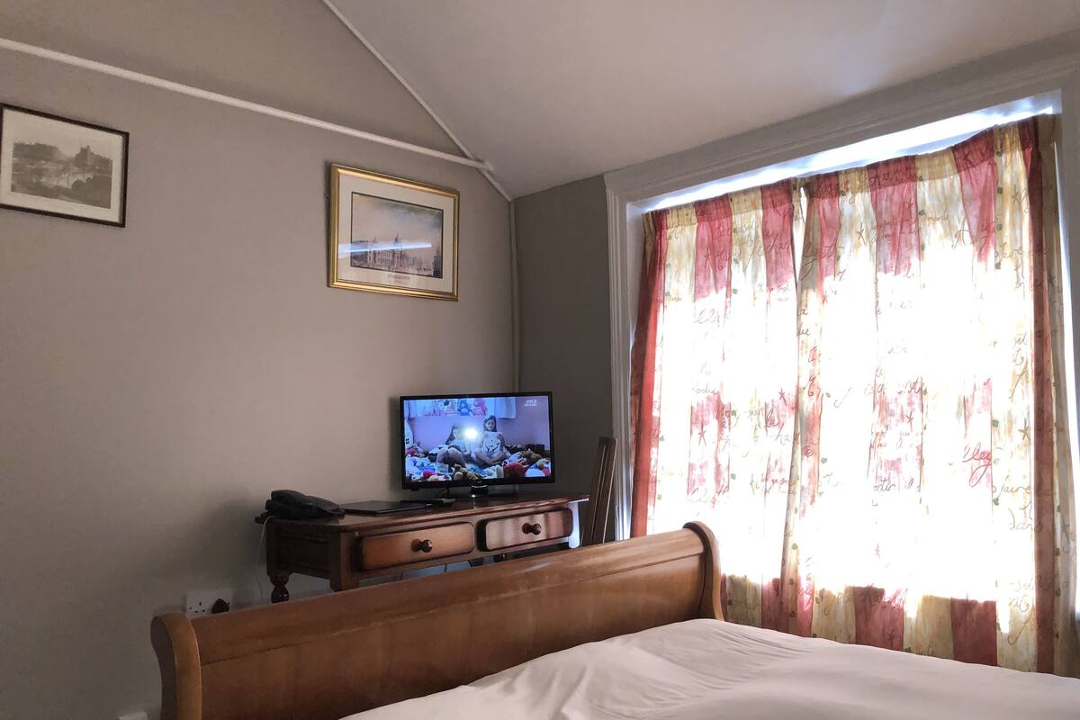 A bedroom with a bed, a television on a wooden dresser, and a window with curtains.