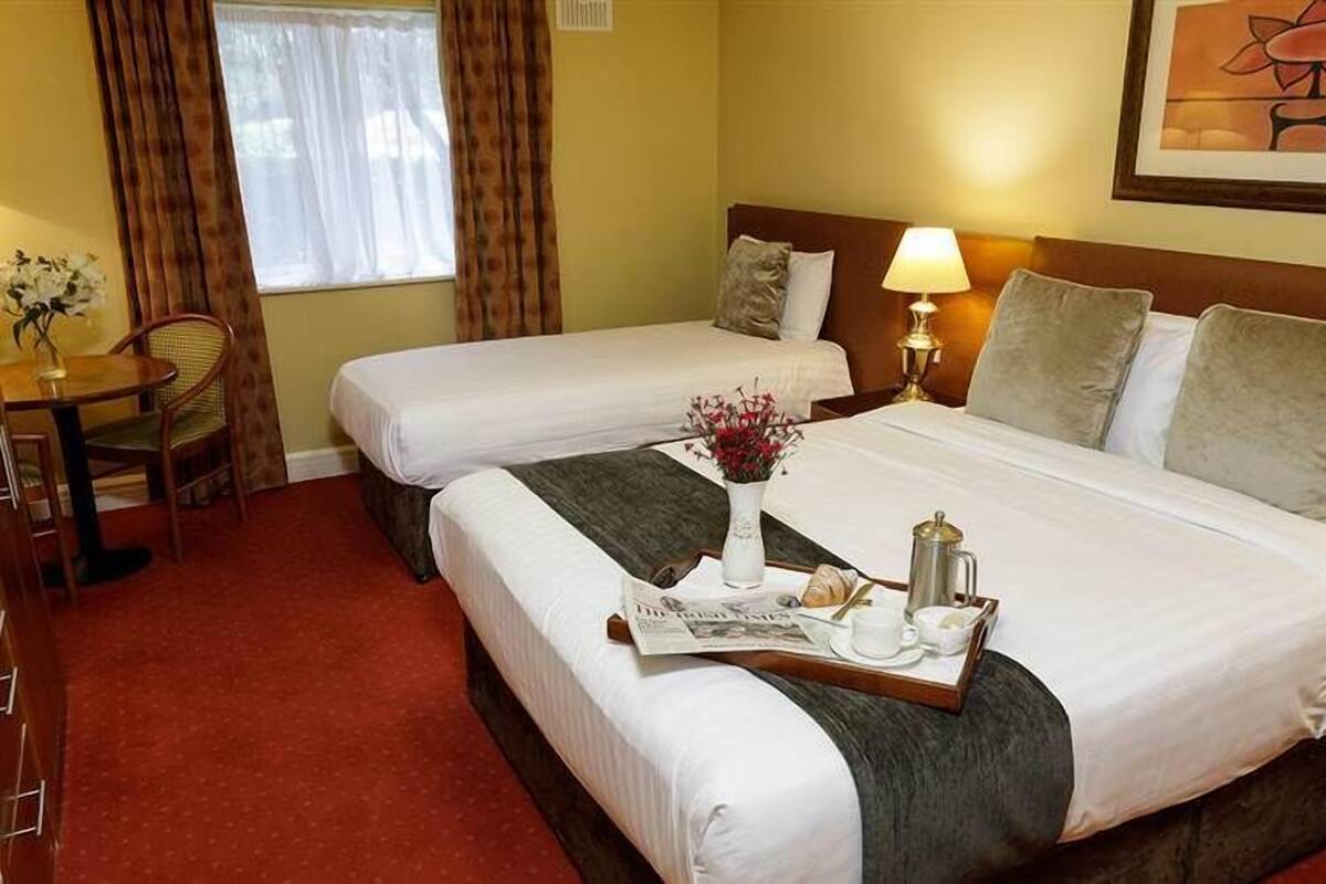 A hotel room with two beds, a small table with a chair, a lamp, and a painting on the wall.