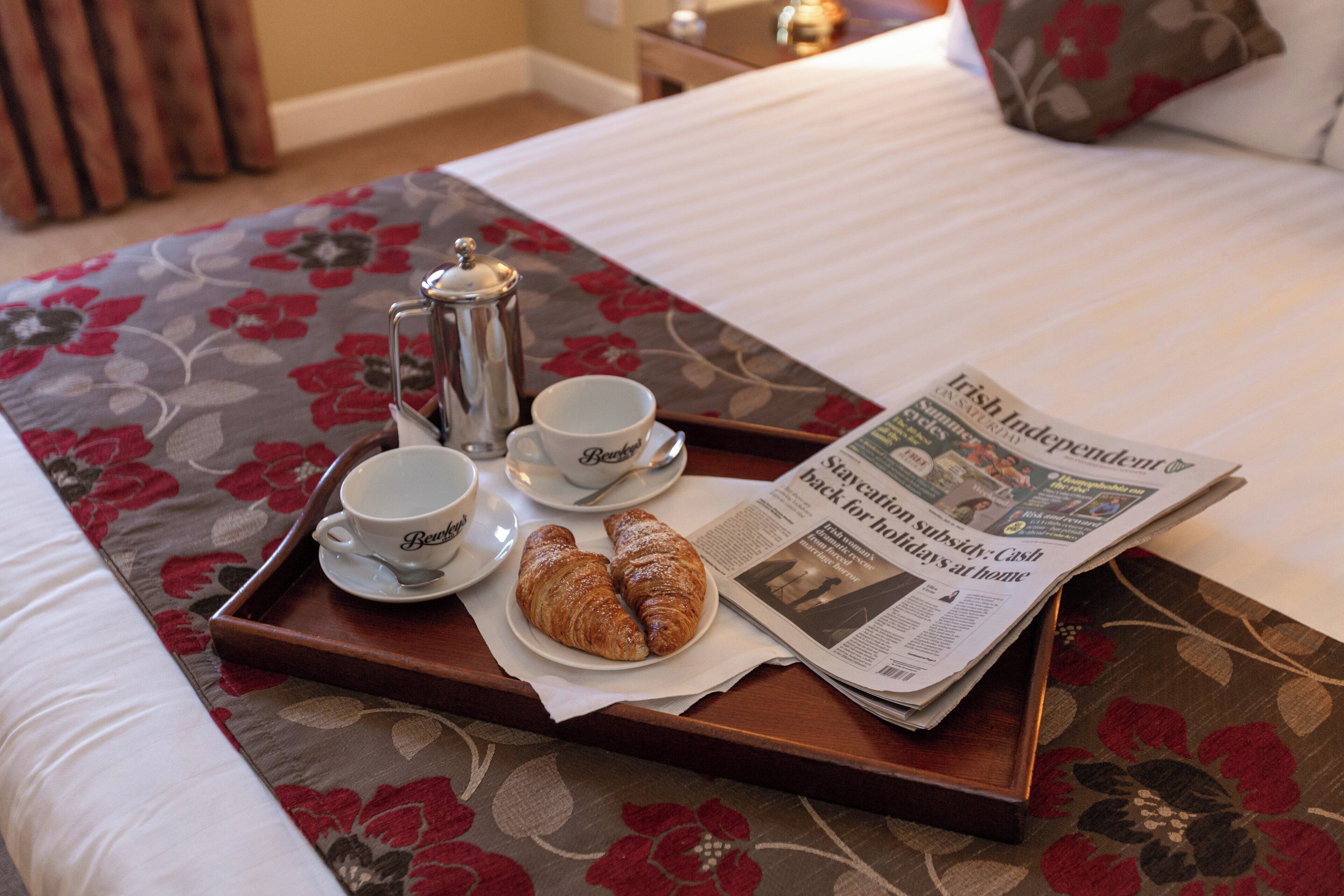 A tray with croissants, coffee cups, and a newspaper on a bed with floral bedding.