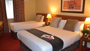 A hotel room with two beds, a wooden headboard, a red carpet, a framed picture on the wall, and a small table with a lamp and flowers. - Hamlet Court Hotel (Johnstown Bridge)