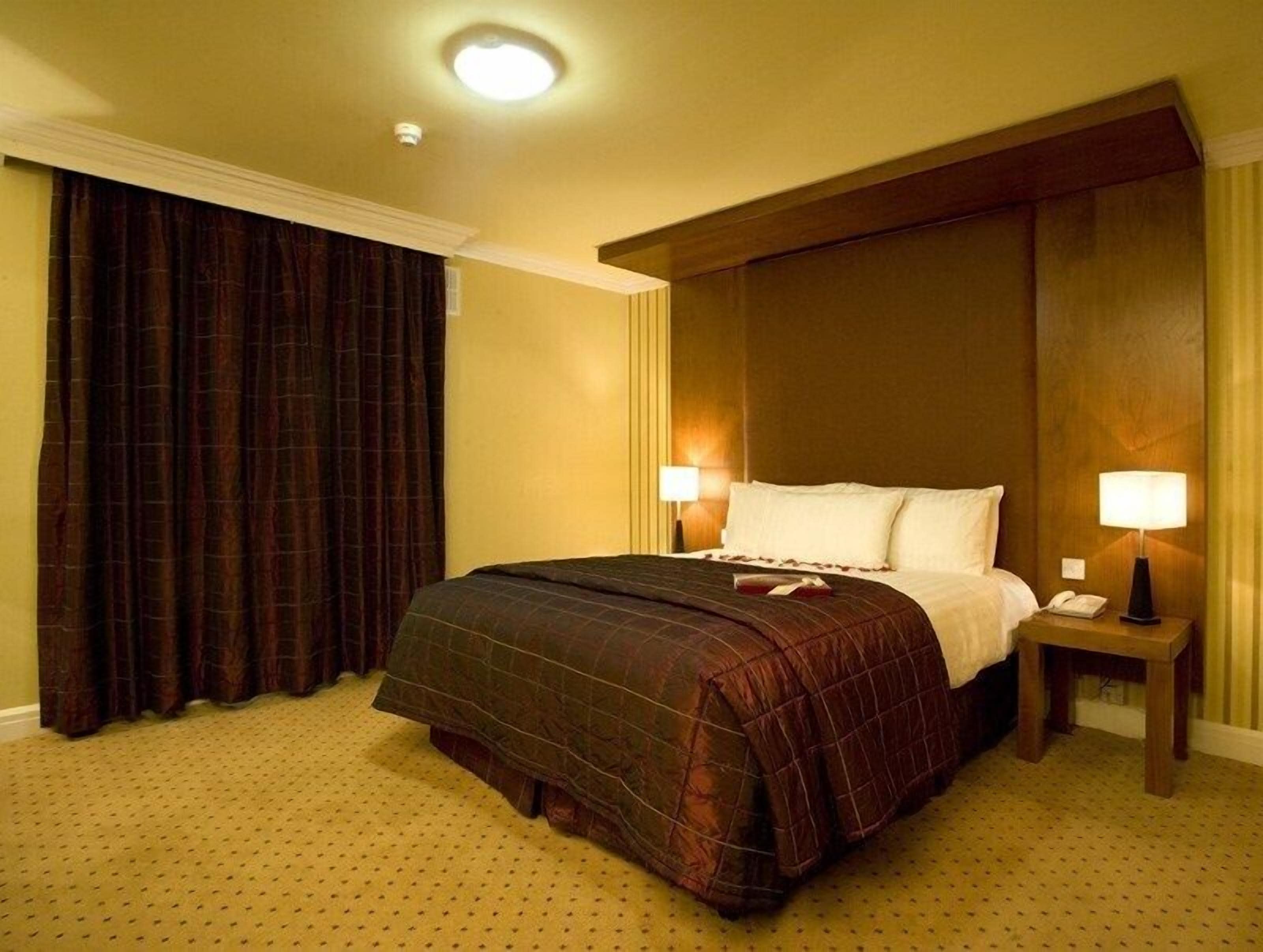 A hotel room with a large bed, two bedside tables with lamps, a nightstand, and a large window with curtains.