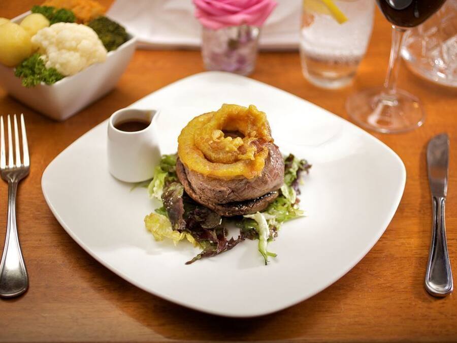 Prato com bife coberto de anéis de cebola, acompanhado de um pequeno recipiente com molho, e uma saladeira ao fundo.