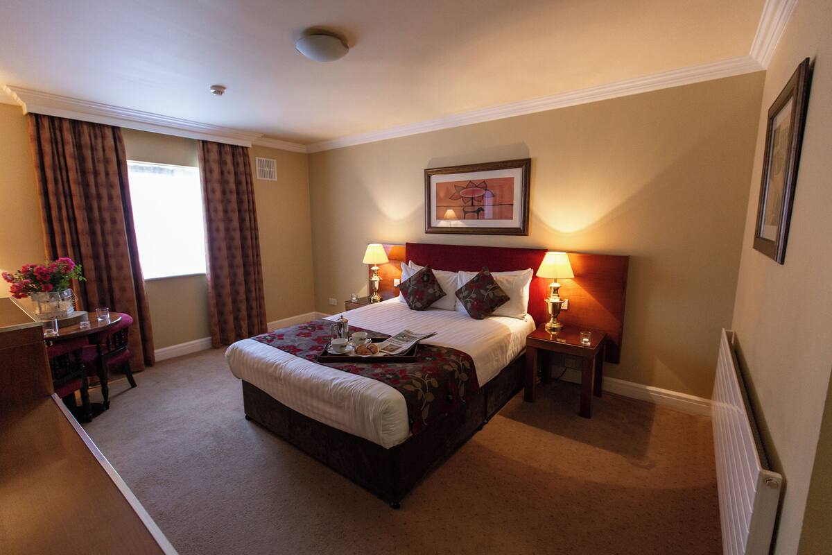 A hotel room with a large bed, two bedside tables with lamps, a small table with a vase and flowers, and a window with curtains.