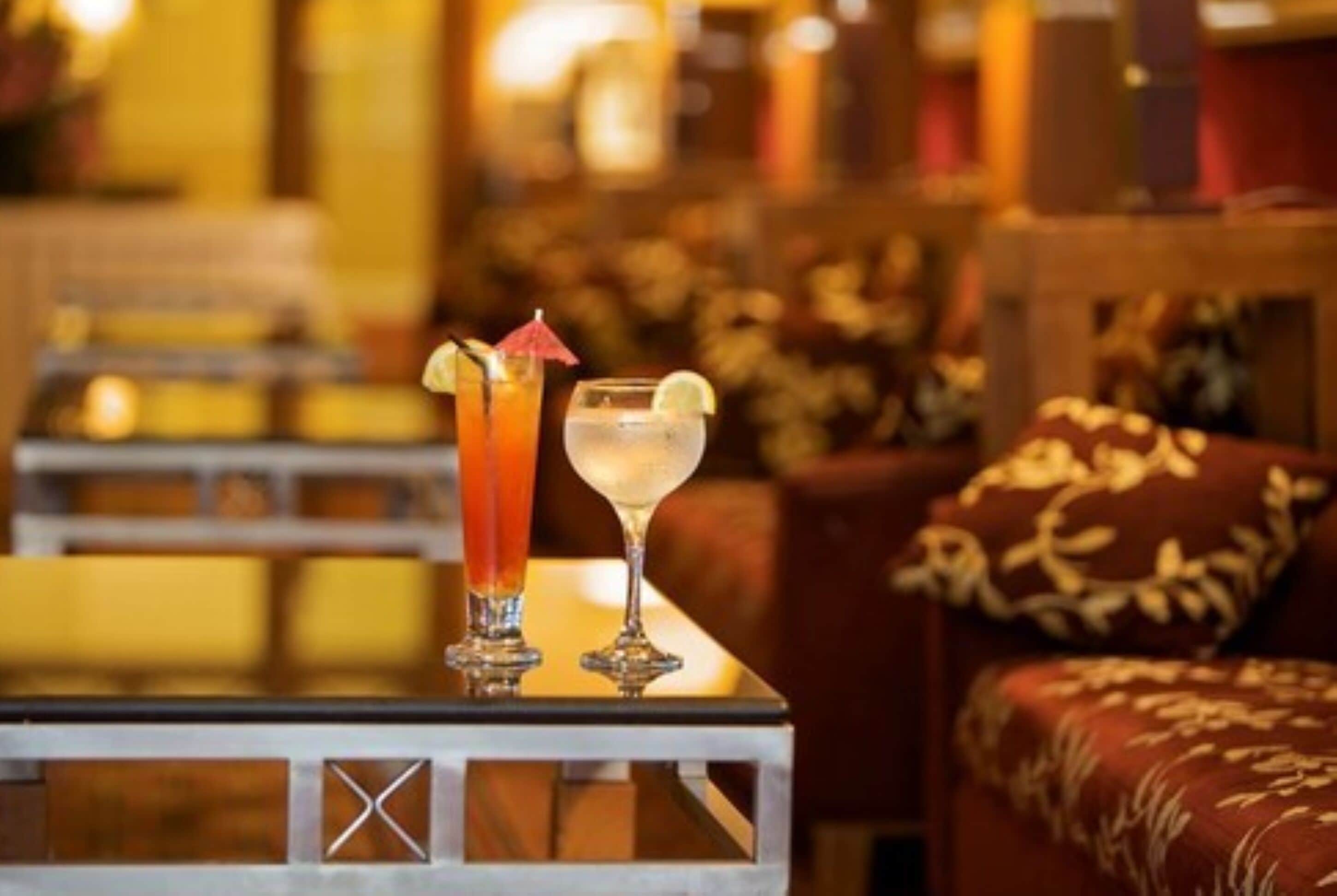 Two cocktails on a table with a patterned cushion in the background.