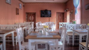 Un comedor con mesas y sillas de madera blanca, una araña y un televisor en la pared.