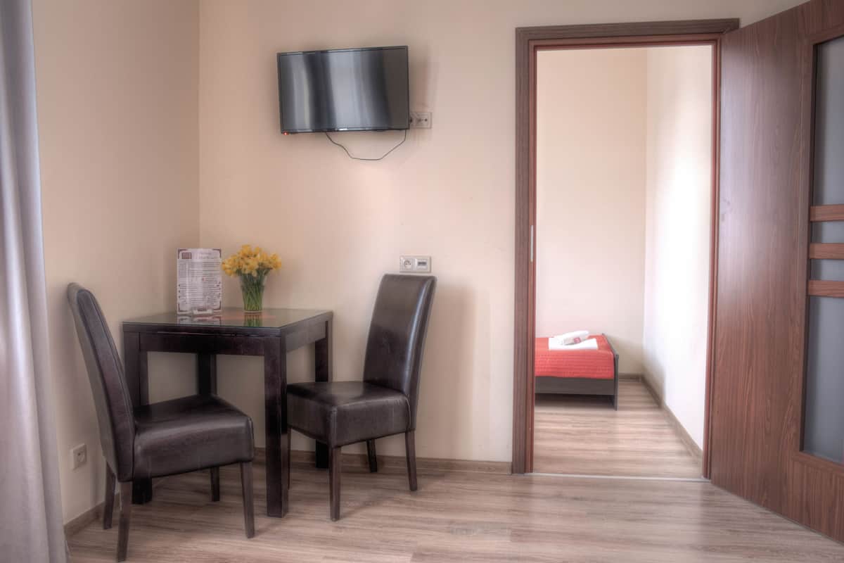 Um quarto pequeno com piso de madeira, uma mesa com duas cadeiras, uma TV fixada na parede e uma cama ao fundo.