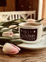 A white mug with "BED ROOMS" text on a saucer, surrounded by pink tulips.