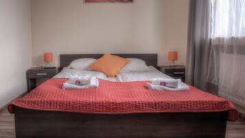 Una cama bien hecha con colcha roja, dos almohadas y dos toallas sobre ella. Hay dos mesitas de noche con lámparas, un cuadro en la pared y una ventana con cortinas.