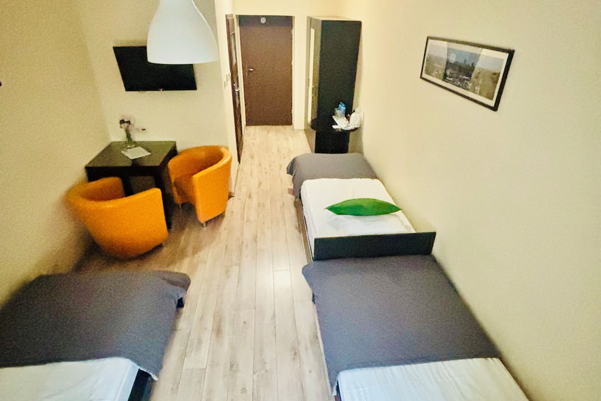 Quarto de hotel com duas camas de solteiro, uma pequena mesa com uma cadeira, uma luminária pendente e um quadro na parede.