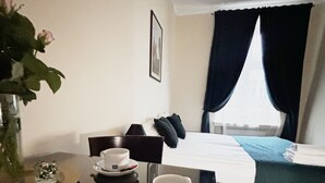 Habitación de hotel con cama, mesita de noche con taza y plato, un florero con flores y ventana con cortinas.