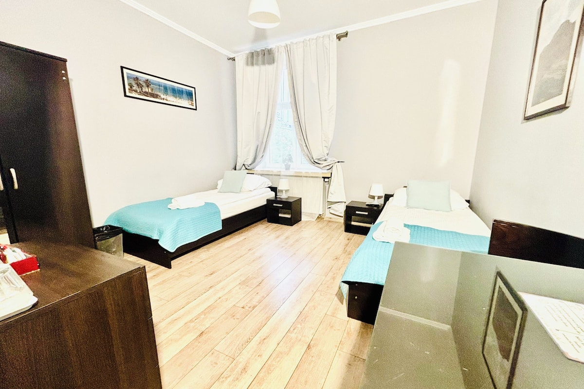 Quarto de hotel com duas camas de solteiro, uma escrivaninha, uma cadeira, janela com cortinas e um quadro na parede.