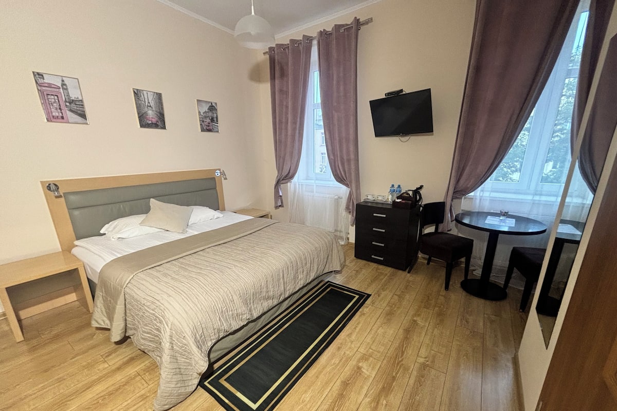 Quarto de hotel com cama, escrivaninha, cadeira, televisão e janela com cortinas.