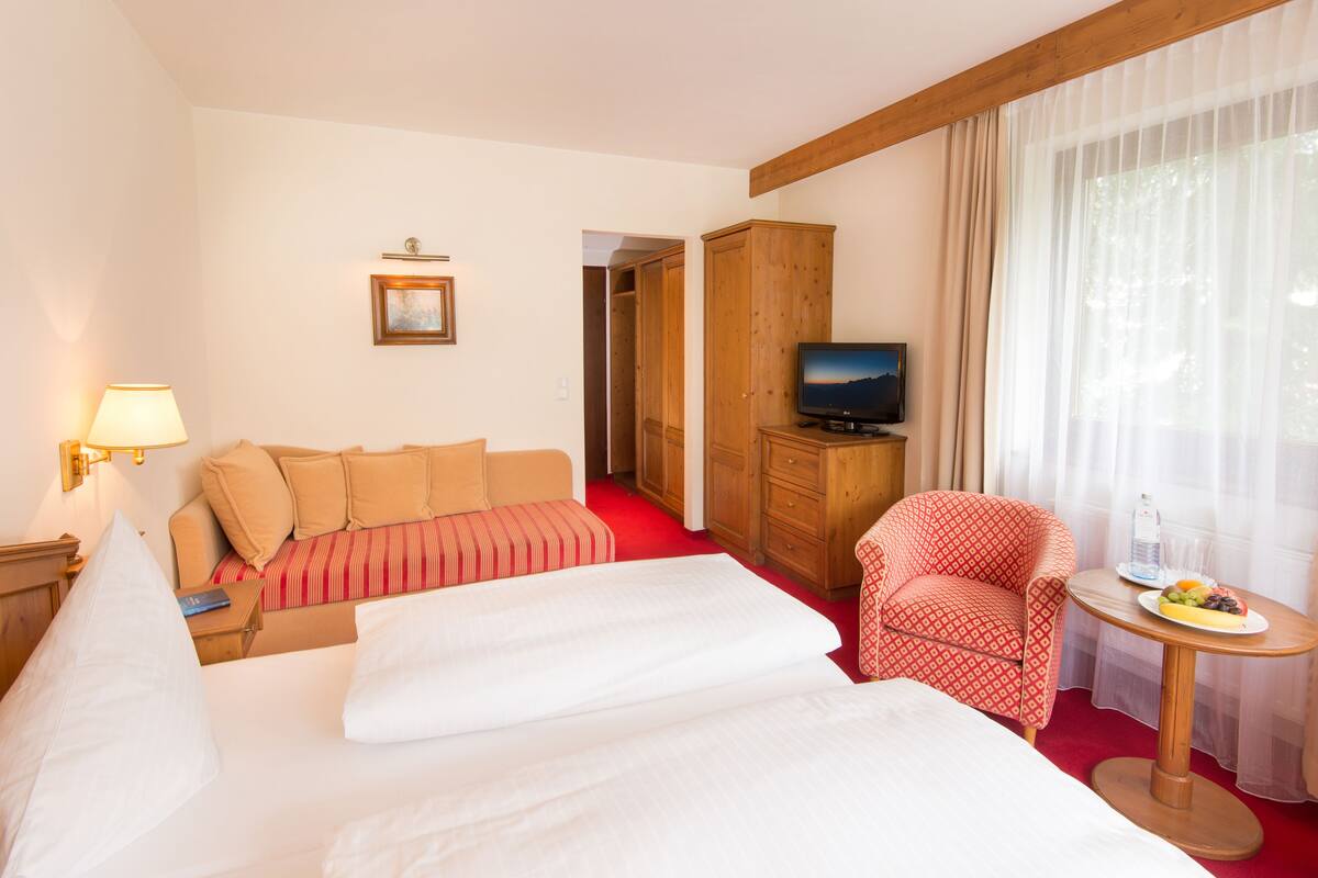 Une chambre d’hôtel avec un lit, une télévision, un fauteuil rouge, une petite table avec des fruits et une armoire en bois.