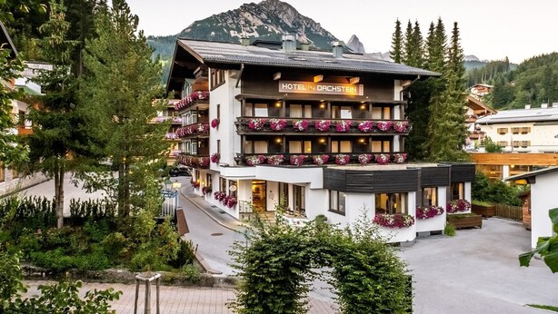 A multi-story hotel with balconies adorned with flowers, set against a mountainous backdrop. - Hotel Dachstein (Filzmoos)