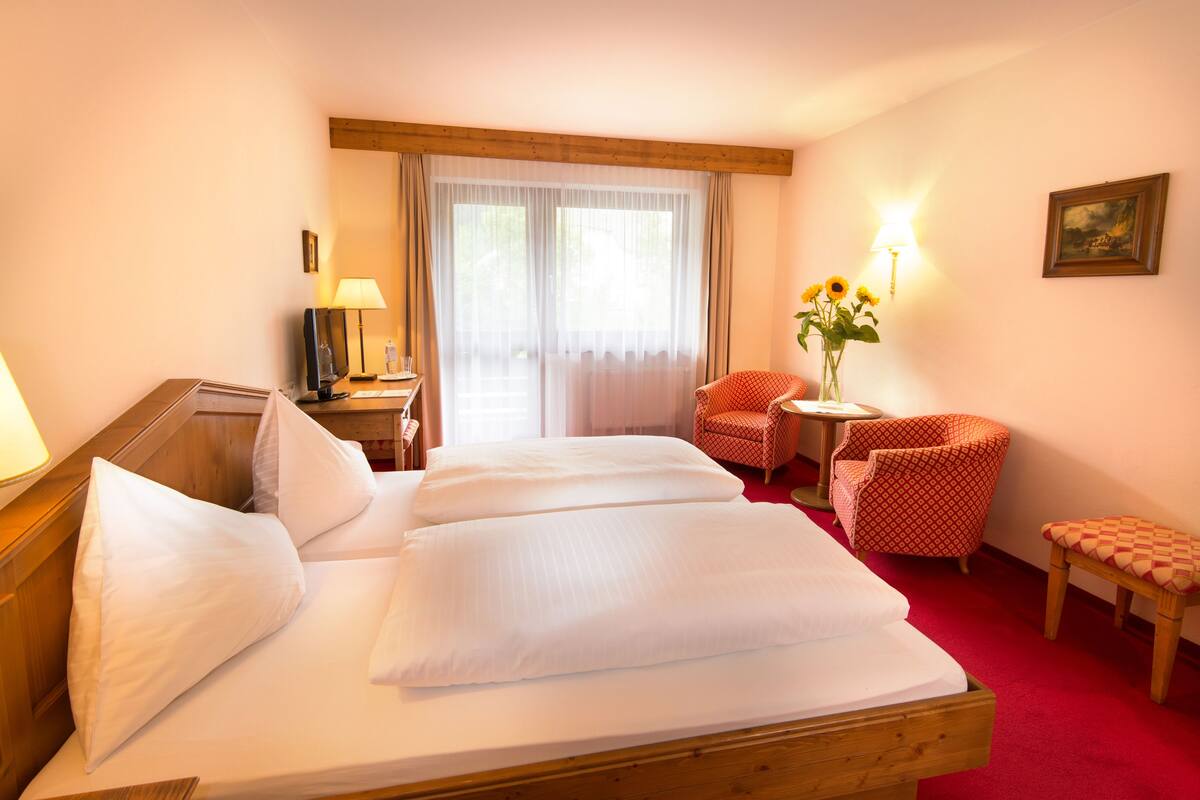 Une chambre d’hôtel avec un lit en bois, un bureau avec une lampe, une chaise, une petite table sur laquelle est posé un vase rempli de tournesols, et une fenêtre ornée de rideaux légers.