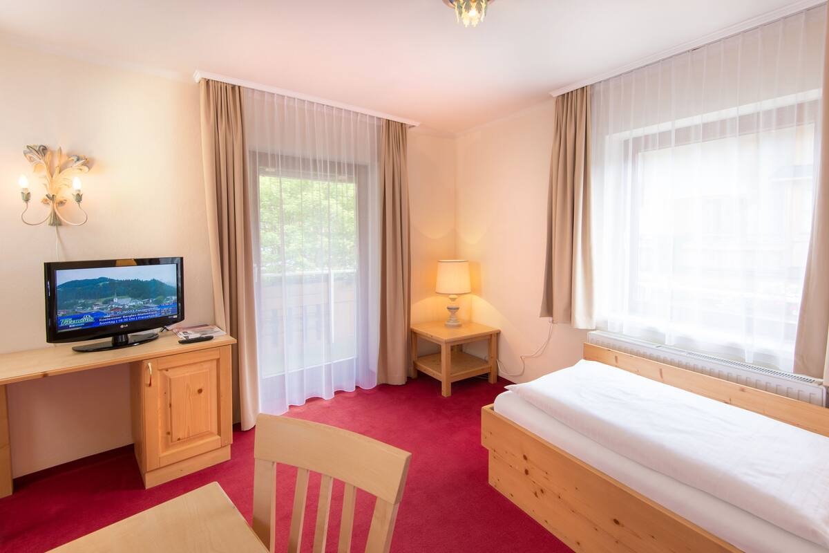 Une chambre d’hôtel avec un bureau en bois, une télévision, un lit, une chaise, une table de chevet avec une lampe et des rideaux légers.