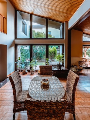 A spacious patio with a round table and wicker chairs, large windows offering a view of the outdoors, and a wooden ceiling.