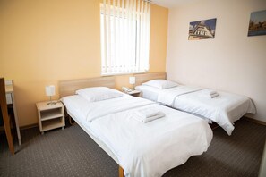 A hotel room with two single beds, a wooden nightstand with a lamp, a window with blinds, and a wall-mounted picture. - Tomas (Šiauliai)
