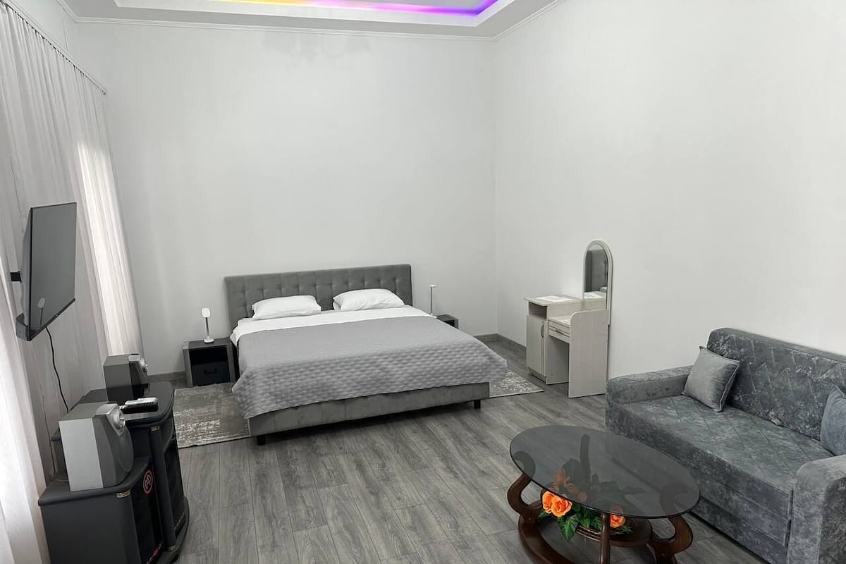 A modern hotel room with a bed, a sofa, a coffee table, a TV, and a desk with a lamp.