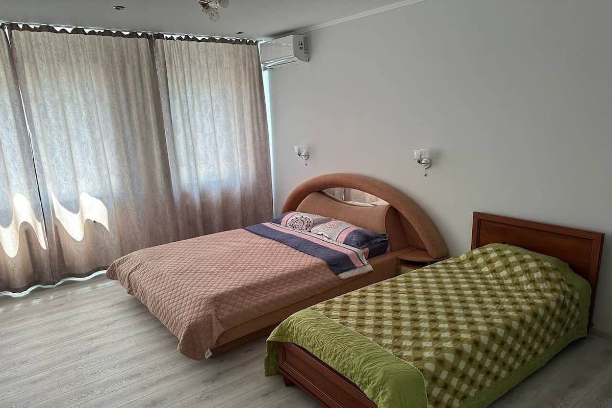 A room with two single beds, a wooden headboard, a window with curtains, and a light switch on the wall.