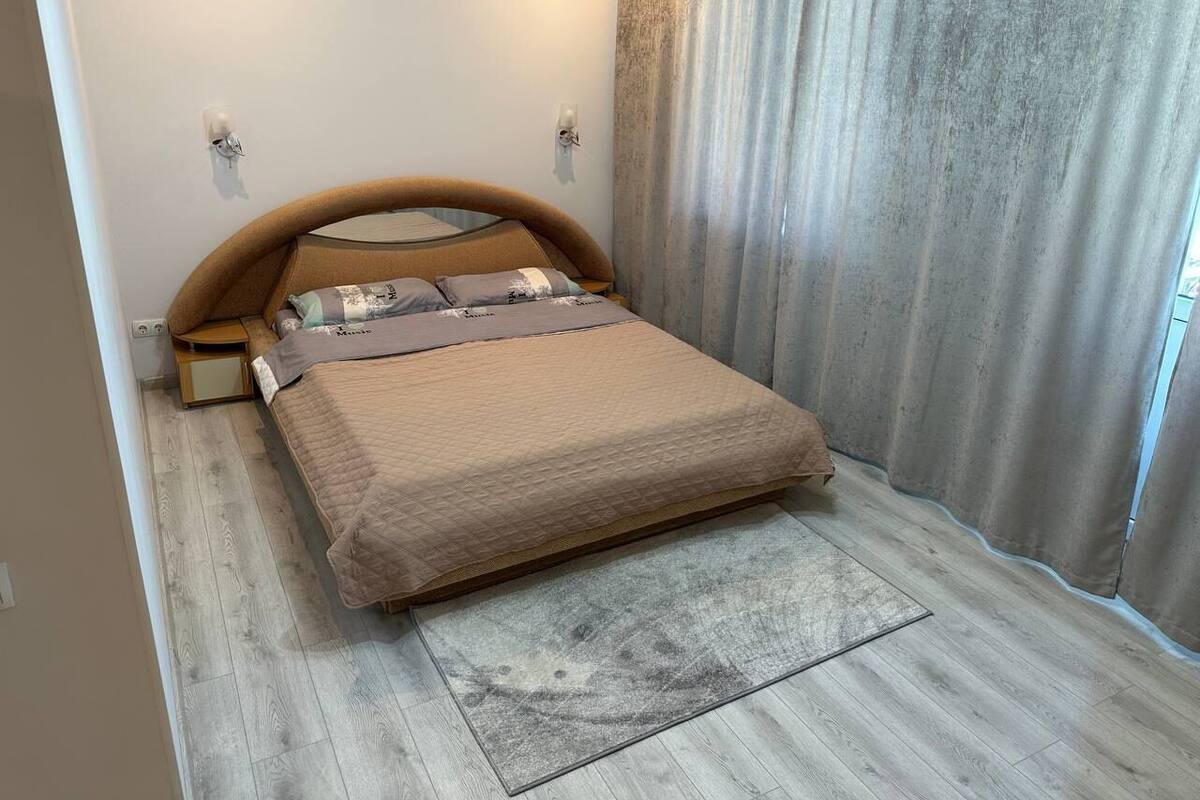 A bedroom with a bed, a nightstand, a wall-mounted light, and a window with curtains.
