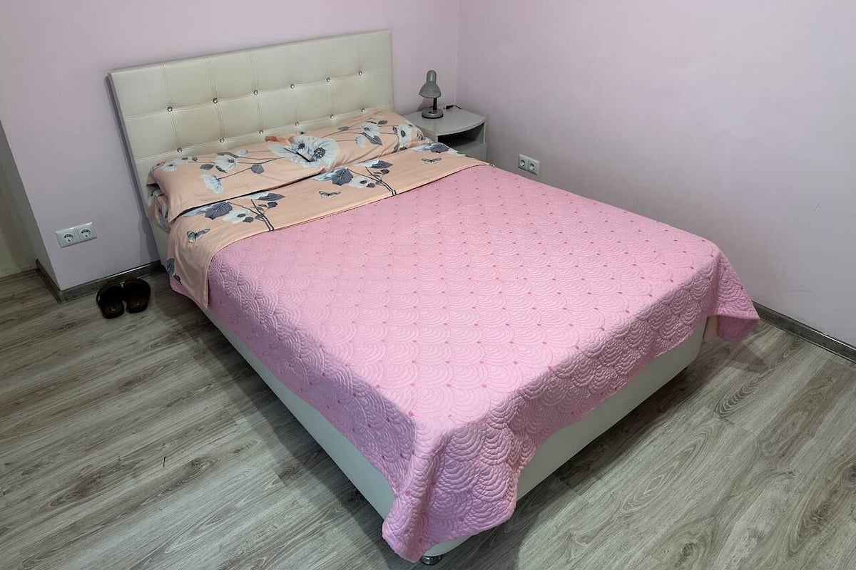 A neatly made bed with a pink quilt and floral-patterned sheets, a bedside lamp, and a pair of shoes on the floor.