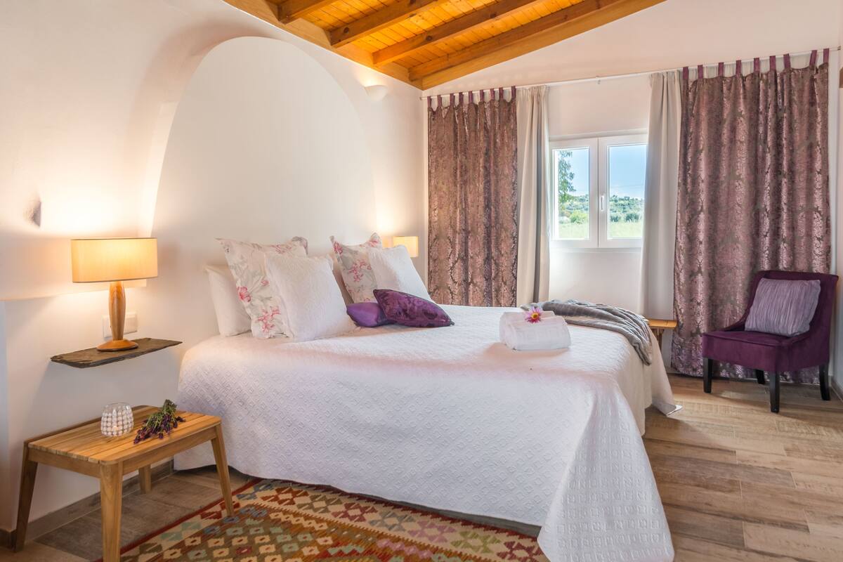 A bedroom with a bed, a nightstand, a chair, and a window with curtains.