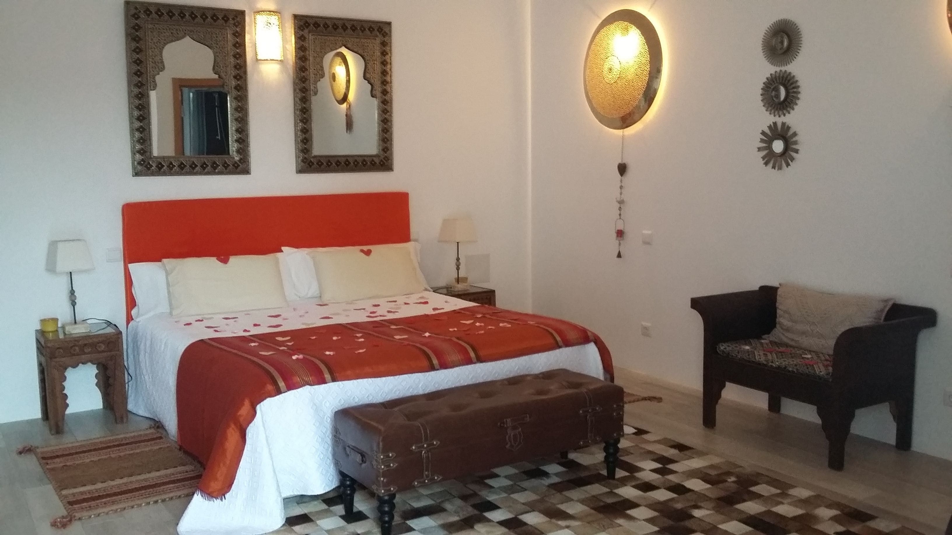 Quarto com cabeceira vermelha, cama grande com colcha estampada, um banquinho, piso xadrez e uma poltrona pequena.