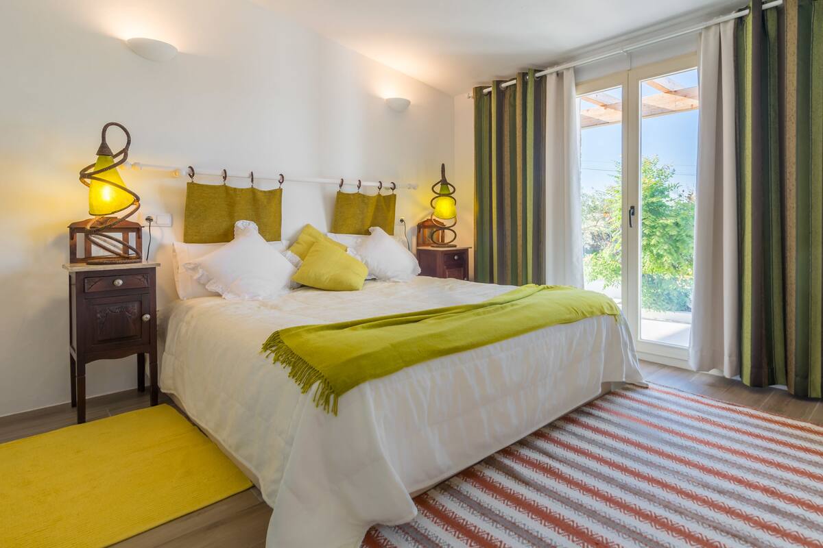 A bedroom with a bed, two bedside tables with lamps, a window with curtains, and a view of greenery outside.