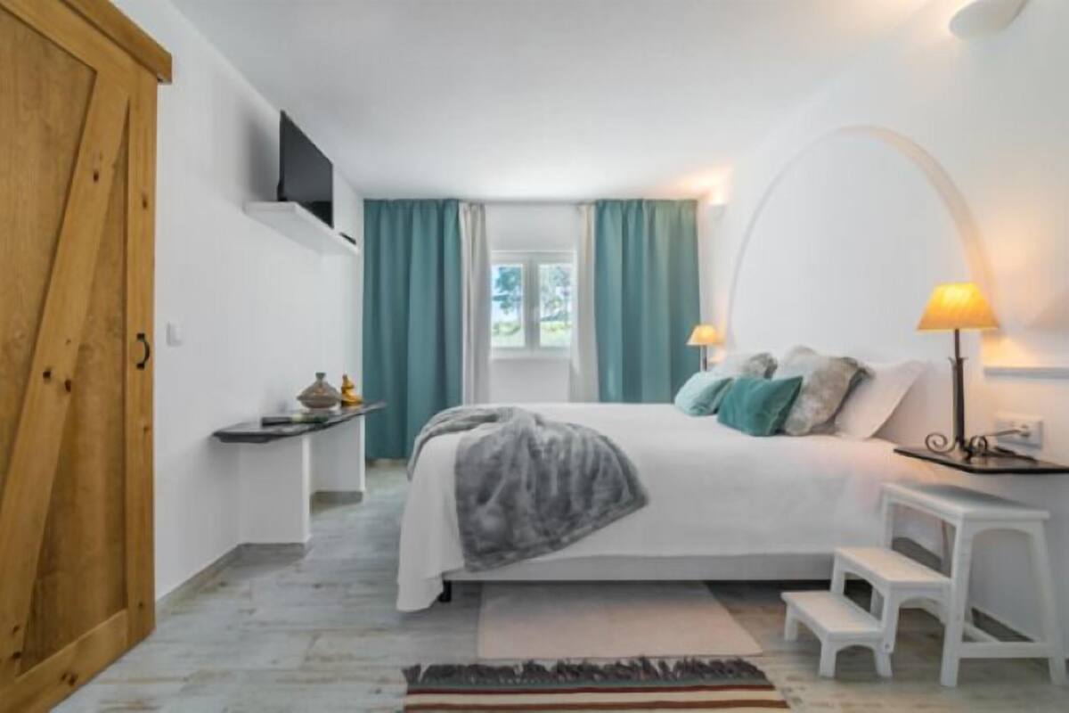A bedroom with a bed, bedside lamps, a small table, a window with curtains, and a wooden door.