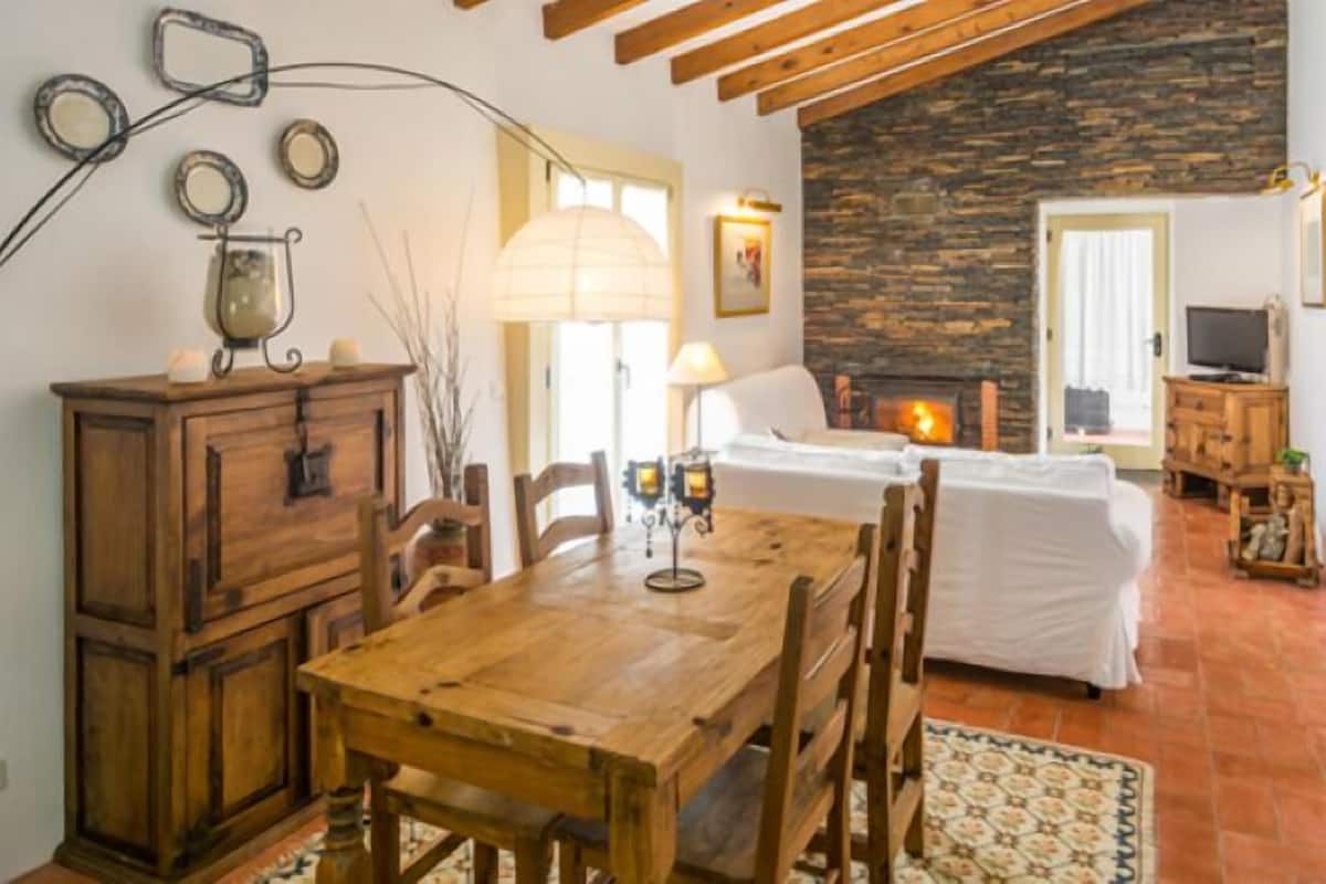 A cozy room with a wooden dining table, a fireplace, a television, and a stone wall.