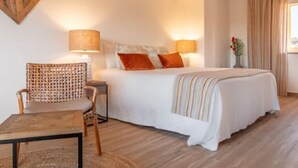 Quarto com cama, duas luminárias de cabeceira, uma cadeira, uma mesinha e um objeto decorativo na parede.