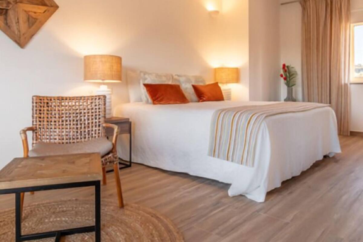 A bedroom with a bed, two bedside lamps, a chair, a small table, and a decorative wall piece.
