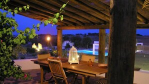Uma pérgola de madeira ao ar livre com uma mesa e cadeiras, um candeeiro em gaiola de pássaro e vista para uma piscina ao anoitecer.